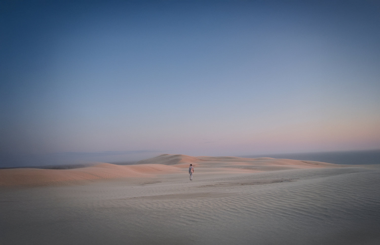 A lone person walks across vast sand dunes under a pastel-hued sky.