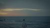 Person in a hoodie stands facing the ocean at dusk, with dark clouds and a fading sunset in the sky.